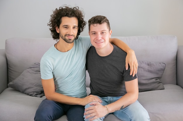 Foto grátis casal homossexual masculino feliz posando em casa, sentados no sofá juntos e se abraçando. vista frontal. conceito de amor e relacionamento