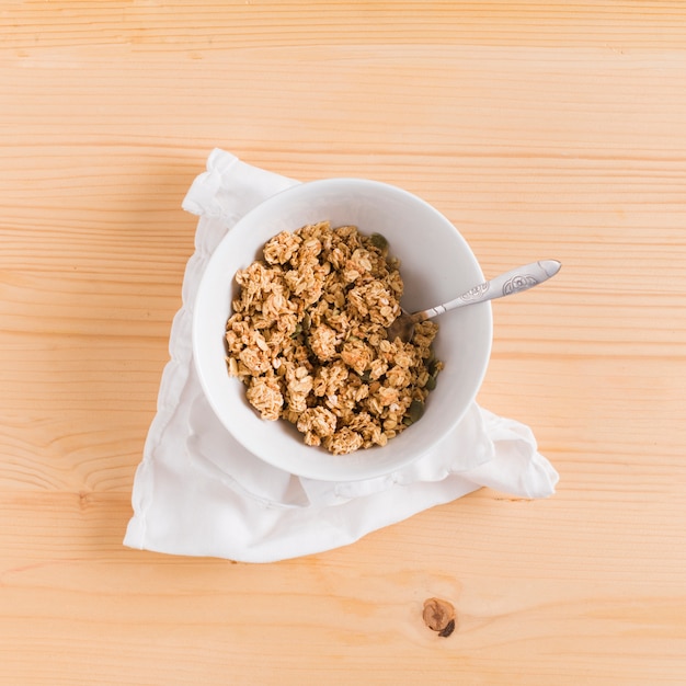 Foto grátis cereais de pequeno-almoço de aveia granola e colher na tigela branca sobre branco guardanapo na mesa de madeira