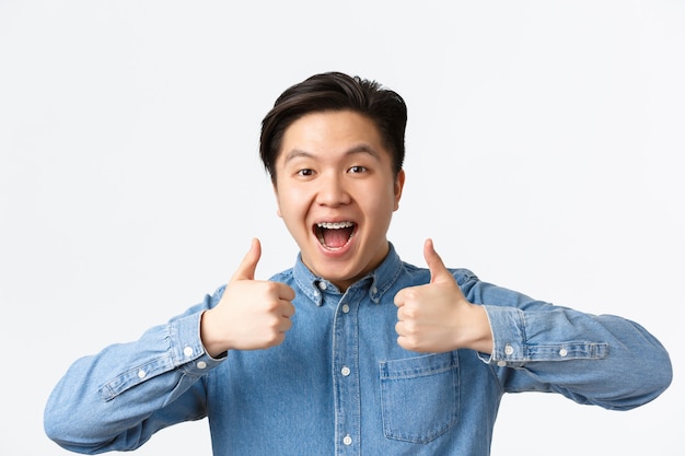 Foto grátis close de uma vitória bem-sucedida, feliz homem asiático com aparelho dentário, sorrindo amplamente e mostrando o polegar para cima em aprovação, elogiando o excelente trabalho, dizendo muito bem, fundo branco de pé satisfeito