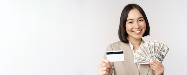 Foto grátis close-up da senhora de escritório de empresária asiática mostrando cartão de crédito e dólares de dinheiro em pé de terno sobre fundo branco