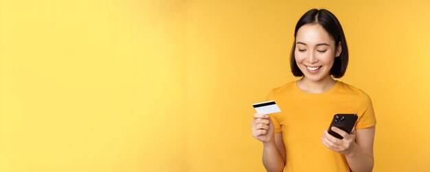 Compras on-line Menina asiática sorridente usando cartão de crédito e aplicativo de celular pagando pedido sem contato no aplicativo de smartphone sobre fundo amarelo