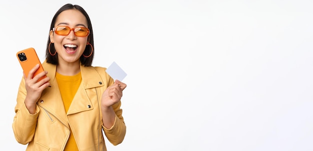 Foto grátis conceito de compras e entrega online garota coreana feliz em roupas elegantes, segurando o cartão de crédito e smartphone rindo e sorrindo em pé sobre fundo branco