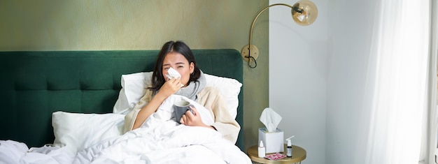 Foto grátis conceito de gripe e vacinação covid garota coreana deitada na cama com resfriado pegando gripe bebendo quente