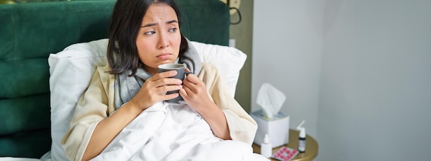 Foto grátis conceito de gripe e vacinação covid garota coreana deitada na cama com resfriado pegando gripe bebendo quente