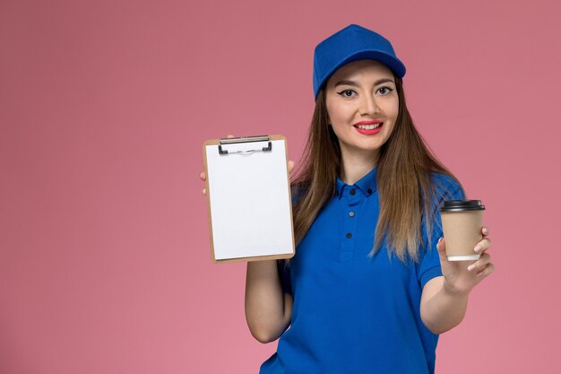 Courier feminino de vista frontal em uniforme azul e capa segurando a xícara de café de entrega e o bloco de notas na secretária rosa