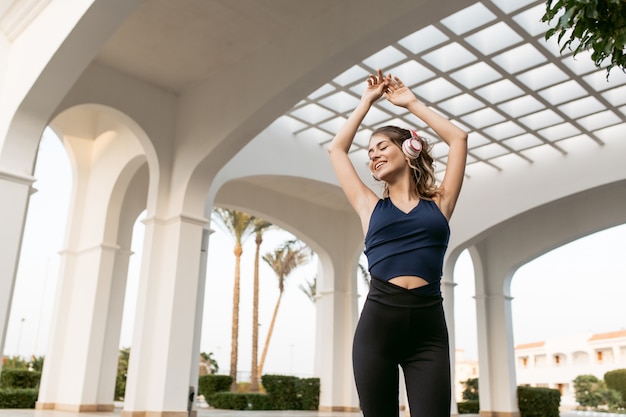 Foto grátis desfrutando de uma música adorável através dos fones de ouvido da desportista alegre positiva, estendendo as mãos acima na arquitetura oriental. modelo fashion, se divertindo ..