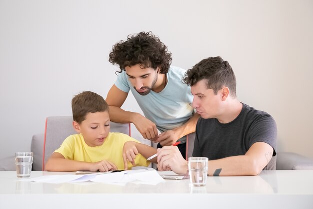 Dois pais ajudando o menino focado com a tarefa escolar de casa, sentado à mesa com papéis, lendo o livro juntos, apontando o dedo na página. Conceito de família e paternidade