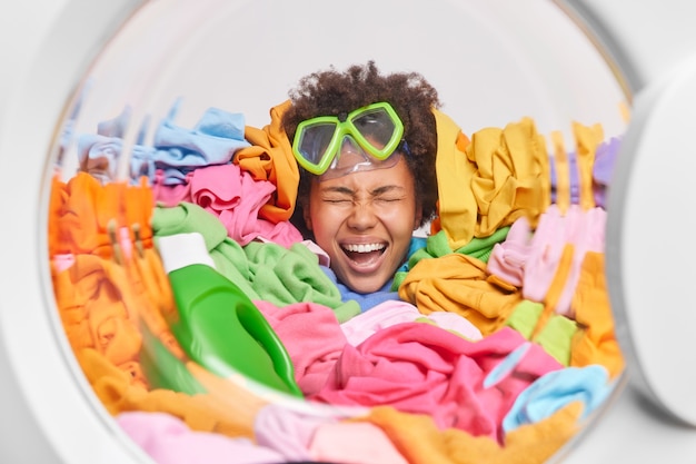 Foto grátis emocional, alegre, feliz, dona de casa afro-americana sobrecarregada com tarefas domésticas tem a cabeça gravada nas tarefas domésticas em um monte de roupas coloridas e usa máscara de mergulho na testa