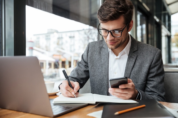 Foto grátis empresário sorridente em óculos, sentado junto à mesa no café com o computador portátil enquanto estiver usando o smartphone e escrevendo algo