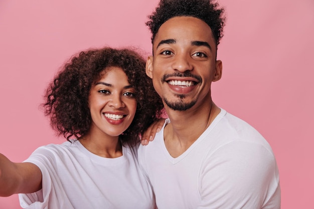 Foto grátis encantadora mulher e homem vestido de camisetas brancas sorrindo e tirando selfie retrato de alegre menina encaracolada e menina de pele escura em tee posando em fundo rosa