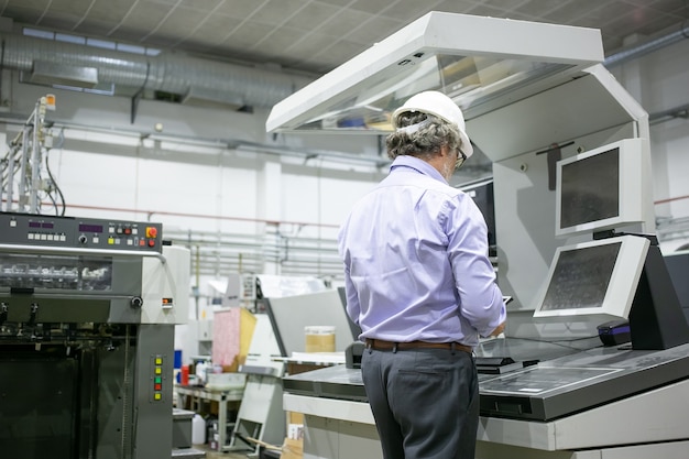 Foto grátis engenheiro de planta masculino de cabelos grisalhos, capacete e óculos, parado na máquina industrial, usando dispositivo digital