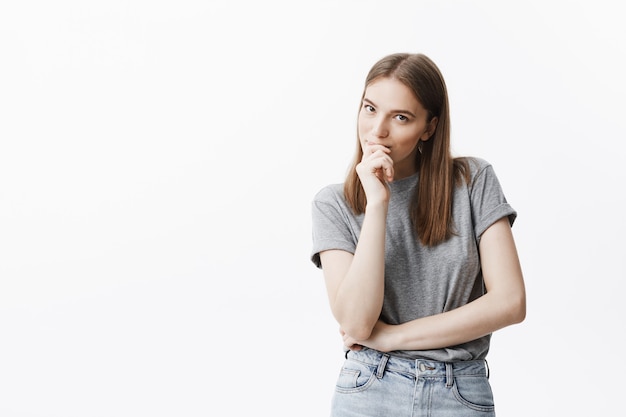 Foto grátis feche o retrato da bela morena caucasiana jovem com cabelos longos em t-shirt cinza e calça jeans com expressão de rosto pensativo, sorrindo, segurando a mão perto da boca