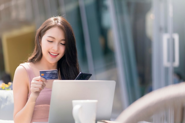 Foto grátis felicidade feminina asiática adulta atraente compras on-line com cartão de crédito de mão de laptop e smartphone para compra de produtos com fundo de construção de jardim conceito de ideias de negócios ao ar livre