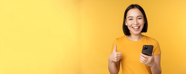 Foto grátis feliz garota asiática sorridente segurando o celular e mostrando os polegares para cima, recomendando o aplicativo no smar