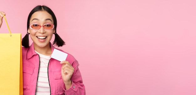 Foto grátis feliz jovem asiática mostrando cartão de crédito para compras segurando saco comprando à venda indo para a loja de pé sobre fundo rosa