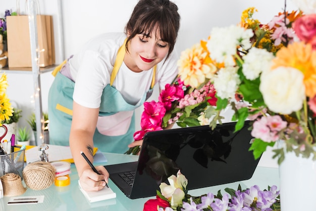 Foto grátis florista feminina escrevendo no bloco de notas com o laptop sobre a mesa