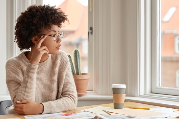 Foto de mulher contemplativa com penteado afro, usa óculos redondos, suéter casual quente