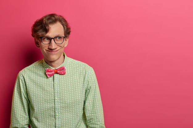 Foto grátis foto de um homem engraçado e alegre usa uma elegante camisa verde e gravata borboleta, óculos transparentes, tem um olhar positivo alegre à parte, planeja algo em mente, isolado na parede rosa, copie o espaço para o texto