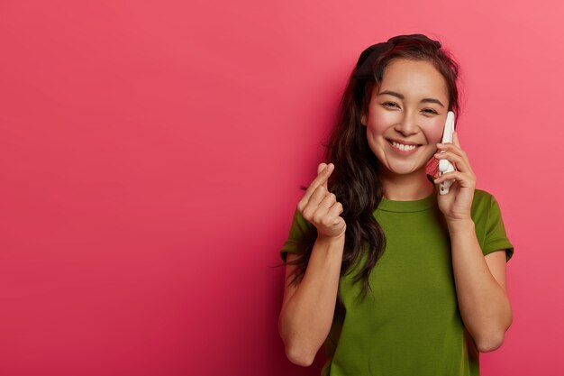 Foto interna de uma garota feminina bonita com um sorriso largo faz um sinal de coreano, expressa afeto enquanto conversa com o namorado ou noivo via celular