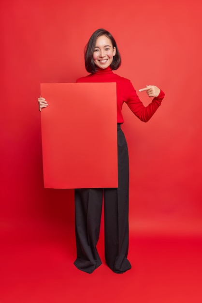 Foto grátis foto vertical de mulher asiática feliz com cabelo escuro e sorriso agradável aponta para papel quadrado para modelo mostra maquete para seu projeto anuncia banner promocional vestido com roupas elegantes poses internas