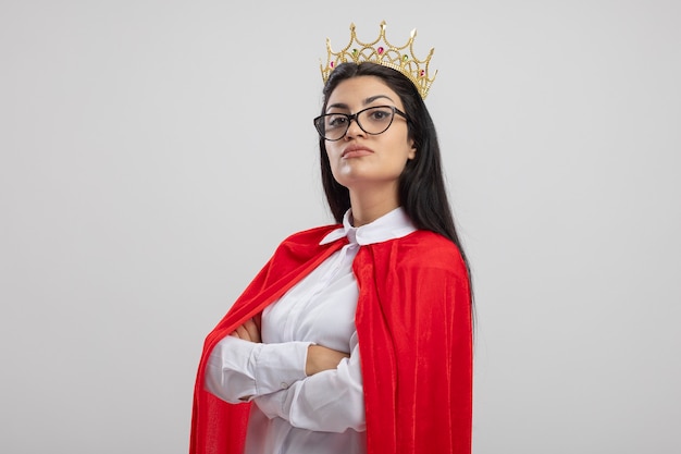 Foto grátis garota jovem super-heroína caucasiana confiante usando óculos e coroa em pé com postura fechada em vista de perfil, olhando para a câmera isolada no fundo branco com espaço de cópia