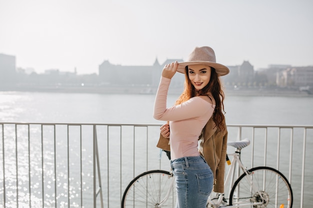 Foto grátis graciosa mulher magra em jeans tocando seu chapéu, parada na ponte na cidade