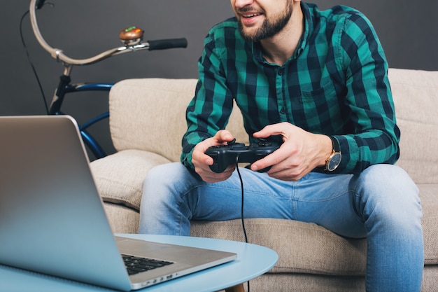 Foto grátis homem barbudo bonito jovem hippie sentado no sofá em casa, jogando videogame no notebook