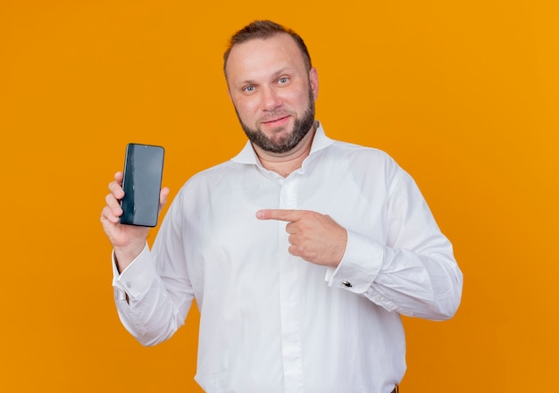 Homem barbudo vestindo camisa branca mostrando smartphone apontando com o dedo para ele e sorrindo em pé sobre a parede laranja