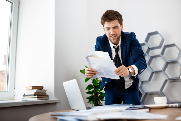 Foto grátis homem de negócios novo irritado que vasculha nos papéis, fundo do escritório.