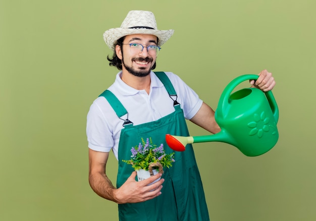 Foto grátis homem jovem jardineiro barbudo vestindo macacão e chapéu segurando um regador e um vaso de planta olhando para a frente sorrindo com uma cara feliz em pé sobre a parede verde