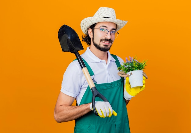 Foto grátis homem jovem jardineiro barbudo vestindo macacão e chapéu segurando uma pá e um vaso de plantas olhando para a frente sorrindo com uma cara feliz em pé sobre a parede laranja