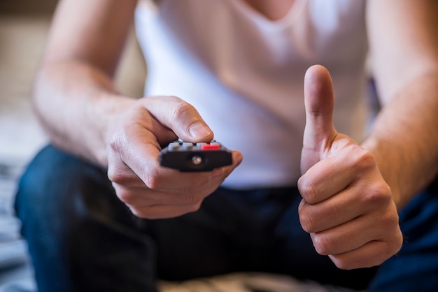 Foto grátis homem segurando um controle remoto de cabo de tv, assistindo tv. estilo de vida, entretenimento, jovens. moda, design e conceito interior. homens com controle remoto mostram polegar para cima