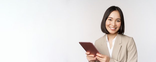 Foto grátis imagem de vendedora de empresária asiática segurando tablet digital e sorrindo trabalhando com gadget em pé de terno sobre fundo branco