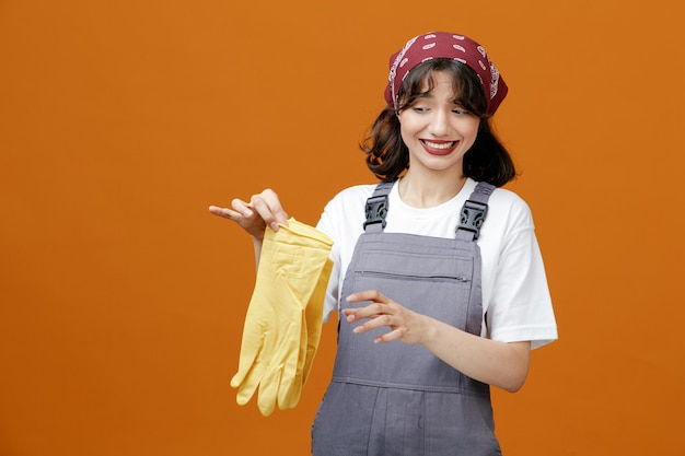 Foto grátis irritada jovem limpador feminino vestindo uniforme e bandana segurando luvas de borracha olhando para eles mantendo a mão no ar isolado em fundo laranja