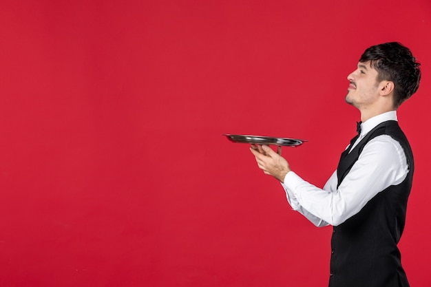 Foto grátis jovem ambicioso garçom em um uniforme amarrando uma borboleta no pescoço segurando uma bandeja sobre fundo vermelho isolado