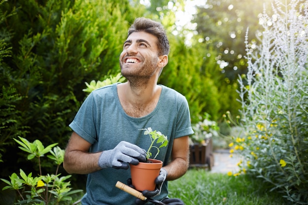 Foto grátis jovem atraente barbudo jardineiro caucasiano com uma camiseta azul e luvas sorrindo, segurando um vaso de flores com broto verde nas mãos, olhando para o lado com expressão de entusiasmo