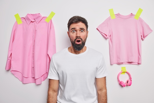 Foto grátis jovem barbudo chocado olha com expressão espantada assustada usa camiseta casual posa contra fundo branco roupas rosa e fones de ouvido colados na parede reage a rumores inesperados