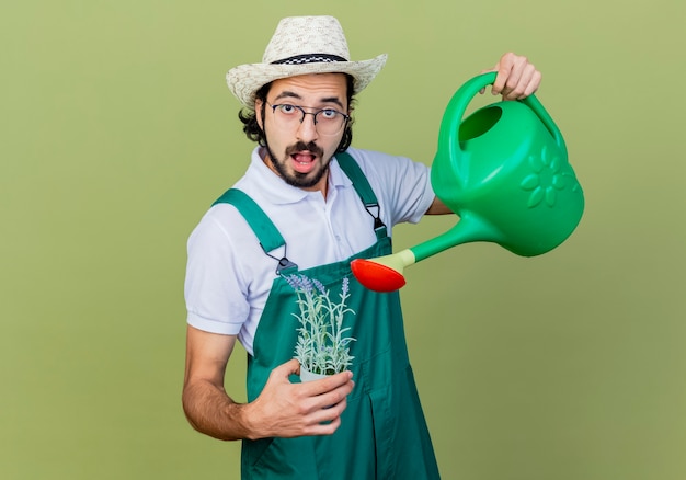 Foto grátis jovem barbudo jardineiro homem vestindo macacão e chapéu segurando um regador e um vaso de planta olhando para a frente surpreso em pé sobre a parede verde claro