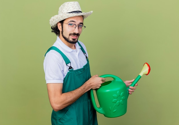 Foto grátis jovem barbudo jardineiro vestindo macacão e chapéu segurando um regador olhando para a frente sorrindo em pé sobre a parede verde claro