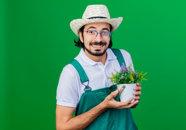 Foto grátis jovem barbudo jardineiro, vestindo macacão e chapéu, segurando um vaso de planta, sorrindo com uma cara feliz em pé sobre um fundo verde