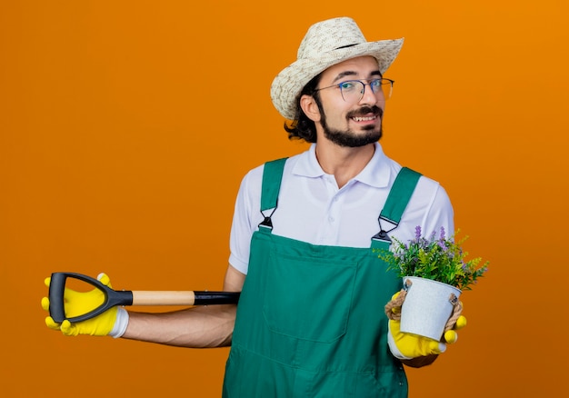 Foto grátis jovem barbudo jardineiro, vestindo macacão e chapéu, segurando uma pá e um vaso de plantas, feliz e positivo, sorrindo