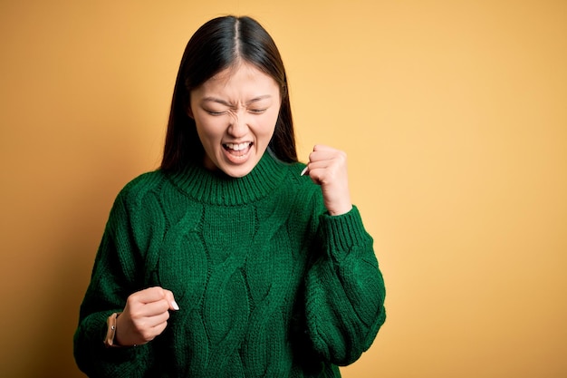 Foto grátis jovem bela mulher asiática vestindo suéter de inverno verde sobre fundo amarelo isolado comemorando surpreso e espantado pelo sucesso com os braços levantados e os olhos fechados conceito de vencedor