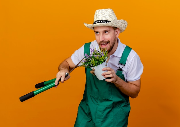 Foto grátis jovem bonito e bonito jardineiro eslavo de uniforme e chapéu segurando uma planta de flor e podadores olhando para cima isolados