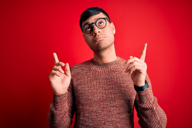 Foto grátis jovem bonito hispânico usando óculos de nerd sobre fundo vermelho apontando para cima parecendo triste e chateado, indicando direção com os dedos infelizes e deprimidos