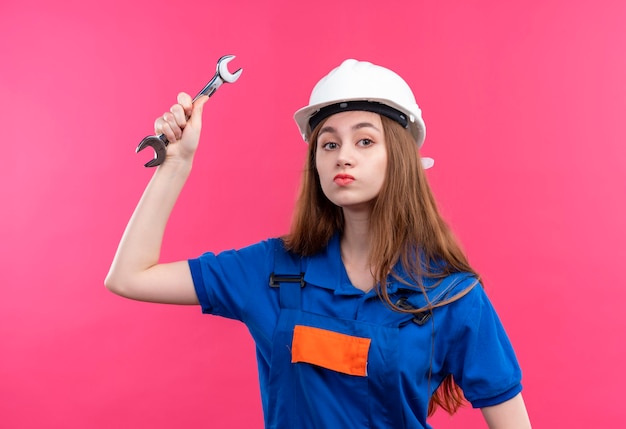 Foto grátis jovem construtora com uniforme de construção e capacete de segurança balançando uma chave inglesa com cara séria em pé sobre a parede rosa