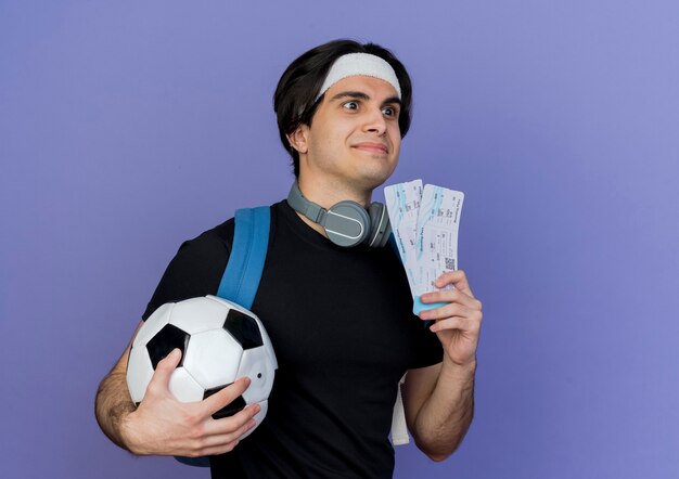 Jovem desportivo vestindo roupas esportivas e bandana com mochila segurando uma bola de futebol e passagens aéreas, olhando de lado sorrindo confuso