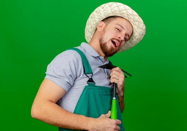 Foto grátis jovem e confiante jardineiro eslavo bonito de uniforme e chapéu segurando um ancinho parecendo fingir cantando usando um ancinho como microfone