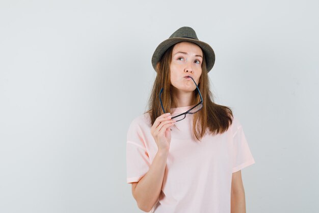 Jovem em t-shirt rosa, chapéu mordendo os óculos e olhando pensativa, vista frontal.