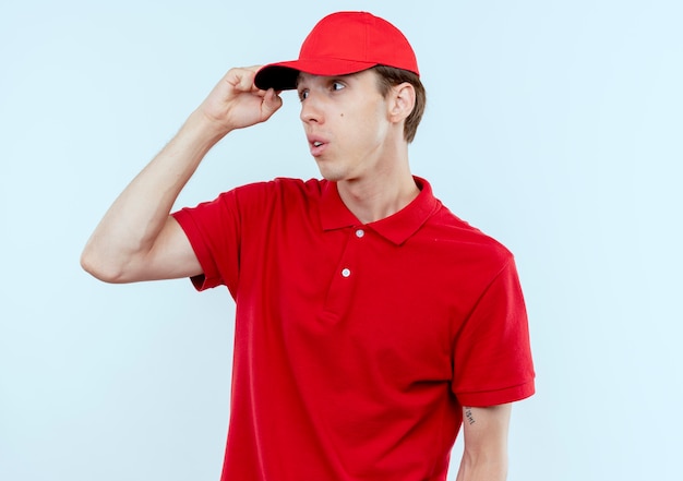 Foto grátis jovem entregador de uniforme vermelho e boné olhando para o lado, confuso com a mão na cabeça em pé sobre uma parede branca