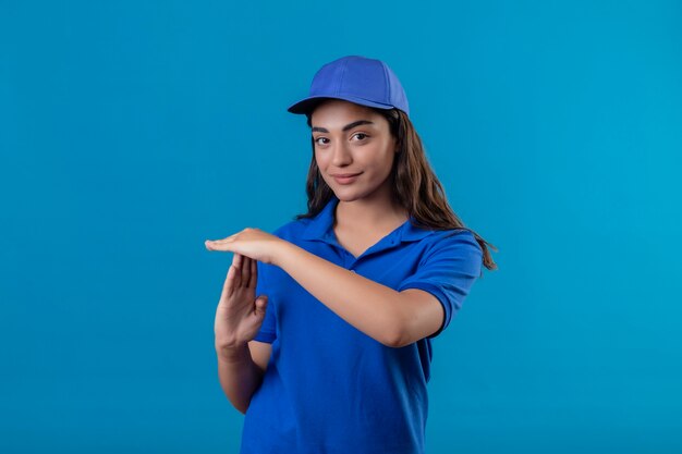 Jovem entregadora de uniforme azul e boné parecendo confiante fazendo gesto de pausa com as mãos em pé sobre um fundo azul